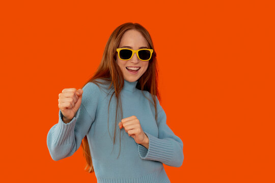 Young woman dances joyfully in colorful sweater and sunglasses on vibrant orange background - Powered by Adobe