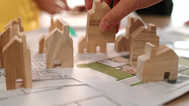 Architect and engineer discuss about building blueprint on table with architectural document, measurement tools scatter around at office, use wooden block for house positioning. Closeup. Delineation.