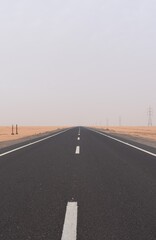 Fototapeta premium the empty asphalt road straight through a desolate desert other side is sandy and barren and high voltage power line tower march along the right side road at Egypt