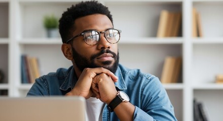 Thoughtful African American man with glasses in deep thought at his desk. Pensive young professional or student planning a project in a modern home office