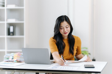 Focused Professional at Work: An Asian professional sits at her desk, penning down notes as she immerses herself in paperwork, enhanced by a modern laptop.