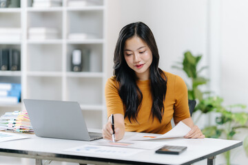 Focused Work: A focused asian woman is engrossed in her work at her desk, the soft light highlighting her concentrated expression as she reviews papers and interacts with her laptop.