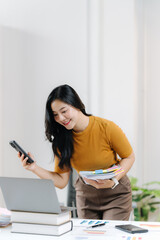 Successful Multitasking: A bright, modern office setting showcases a young professional woman who effortlessly manages several tasks at once, combining the use of smartphone, and computer.