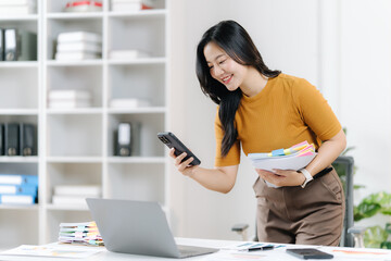Focused Businesswoman At Her Desk: An Asian businesswoman, radiant and dedicated, is fully engrossed in her work at her modern workstation, with a smartphone in one hand and files in the other.
