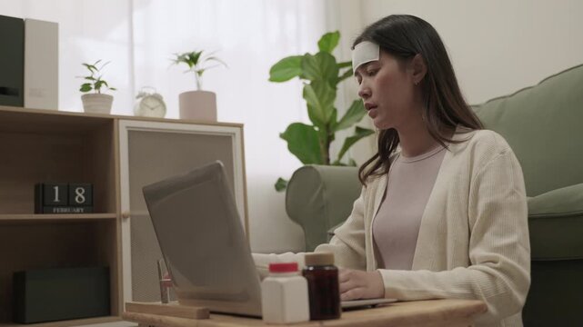 Working from home concept. Young Asian woman employee is symptoms sick and has a headache from the flu use fever cooling gel patch and sitting using laptop computer for working in living room at home.