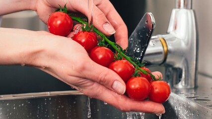 Hands washing a vine of ripe cherry tomatoes under a kitchen faucet