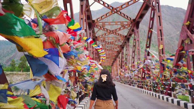 Young woman walking at iron suspension bridge with many Buddhist flags waving in wind. Tourist exploring remote Himalayas. Colorful Tibetan prayer flags on bridge. Tourism background.