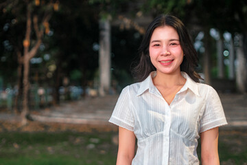 Young woman smiling outdoors in a park setting during golden hour with soft sunlight illuminating her face and background trees creating a serene atmosphere