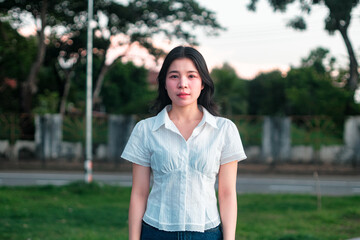 Young woman standing outdoors in casual attire against a backdrop of trees and a soft evening sky, natural light creating a serene atmosphere