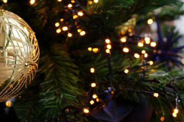 Golden bauble with intricate glitter design hangs on a Christmas tree. In the background are blurred green pine needles and small warm white fairy lights visible, creating a festive glow.