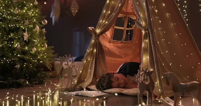 Child sleeping in a cozy teepee near Christmas tree surrounded by wrapped Christmas gifts and twinkle lights