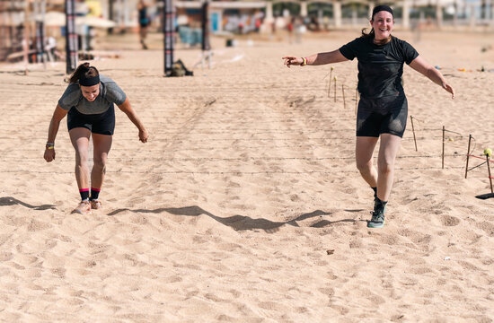 Two joyful female athletes complete the barbed wire crawling section of a beach obstacle course.