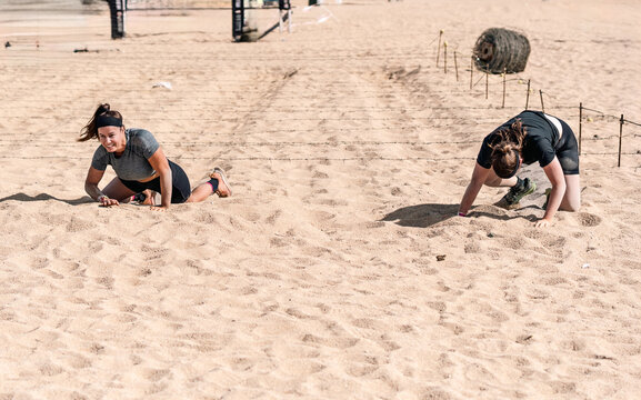 Two determined female athletes emerge from under a barbed wire fence after crawling on the sand in an outdoor obstacle course at the beach.
