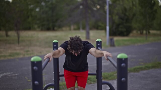 A focused young man with curly hair stretches and strengthens his core on parallel bars in an outdoor gym. He performs a dedicated calisthenics workout, showcasing strength and commitment to fitness.