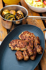 Gebratene Bifteiki (Fleischlaibchen) und Cevapcici auf grauer Servierplatte auf Holztisch, dahinter gegrillte Zucchini, Aubergine und Kartoffeln in Edelstahltopf