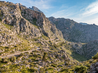 Serpentine road to Sa Calobra, Mallorca, winding mountain road snakes through rugged, rocky terrain with green vegetation, set against a backdrop of towering peaks under a clear blue sky, Majorca