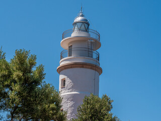 Far del Cap Gros, Port de Soller, Mallorca white lighthouse with a metal railing stands against a clear blue sky, surrounded by lush green trees, Majorca