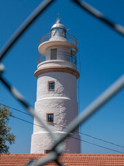 Far del Cap Gros, Mallorca lighthouse Port de Soller, with a white cylindrical tower and a metal railing stands under a clear blue sky, viewed through a chain-link fence, Majorca