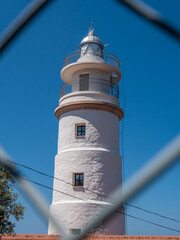 Far del Cap Gros, Port de Soller, Mallorca white lighthouse with a metal railing and a dome on top stands against a clear blue sky, viewed through a chain-link fence, Majorca