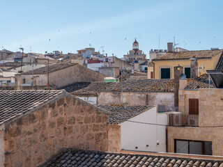 Alcudia, Mallorca picturesque view of an old town with terracotta-tiled roofs, a bell tower, and scattered buildings under a clear blue sky, Majorca