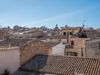 Alcudia, Mallorca, picturesque view of a historic town with terracotta-tiled roofs, nestled against a clear blue sky, with a distant church tower visible in the background, Majorca