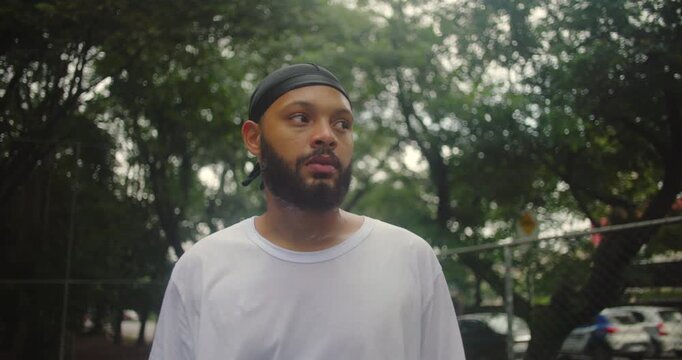 African descent man wearing durag looking away thoughtfully on outdoor basketball court, showing calm determination, introspection, and quiet confidence in an urban athletic setting