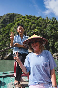 Happy woman wearing traditional conical hat and boat operator making peace signs while exploring the beautiful karst landscapes of ha long bay, vietnam