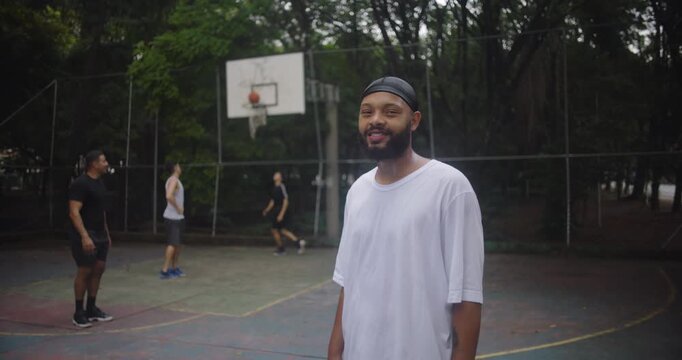 African descent man wearing durag looking calm and confident on basketball court with friends playing behind, representing focus, composure, and strength in teamwork and friendship