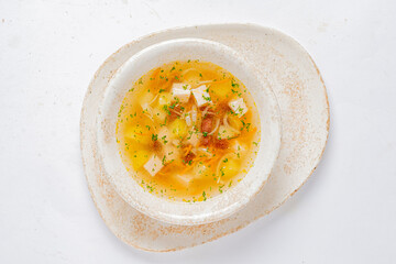 A bowl of yellow vegetable soup sits on a white plate. Cubes of tofu and herbs adorn the soup