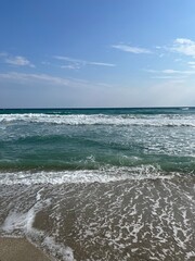 Fototapeta premium Turquoise Waves and White Foam Rolling onto the Sandy Shore Under a Bright Blue Sky