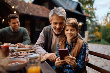 Happy boy and his grandfather using cell phone during family Thanksgiving celebration on patio.