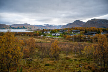 Sortland, Norway, picturesque village nestled among autumn-colored trees, with a serene lake and distant mountains under a cloudy sky, wide angle shot