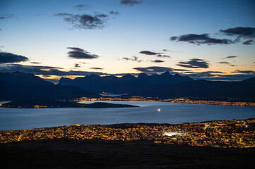 A stunning cityscape of Tromso, Norway with illuminated buildings overlooks a serene lake at twilight, with majestic mountains in the background, at the top of Storsteinen