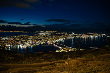 A breathtaking night view of Tromso, Norway illuminated by lights, with a harbor and distant mountains under a dark blue sky, view from Storsteinen
