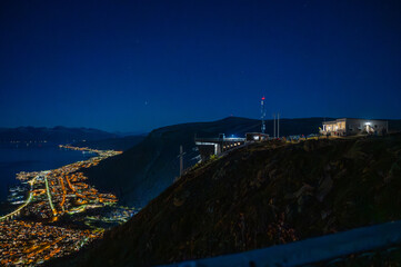 A stunning night view of Tromso, Norway nestled between mountains, with lights illuminating the landscape and a building on a hilltop, at the top of Storsteinen