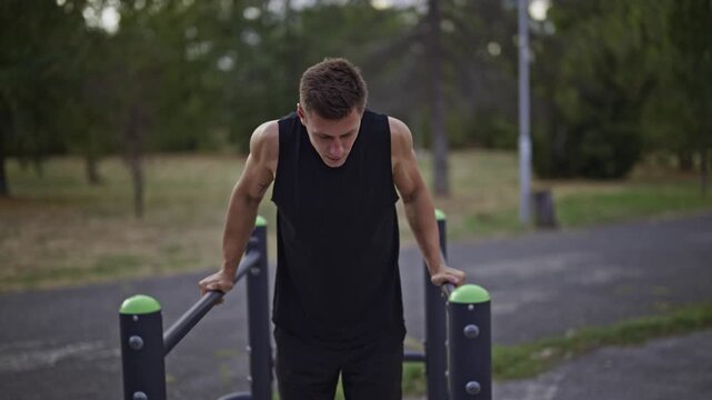 A determined male athlete performs intense dips on parallel bars at an outdoor park gym. He is focused on his fitness routine, showcasing strength and dedication to exercise.