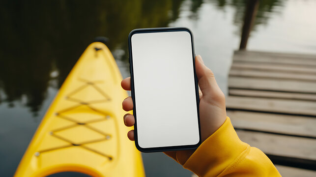 At the lakeside dock, a person holds a blank smartphone next to a yellow kayak, offering a moment for reflection or a digital connection with nature.