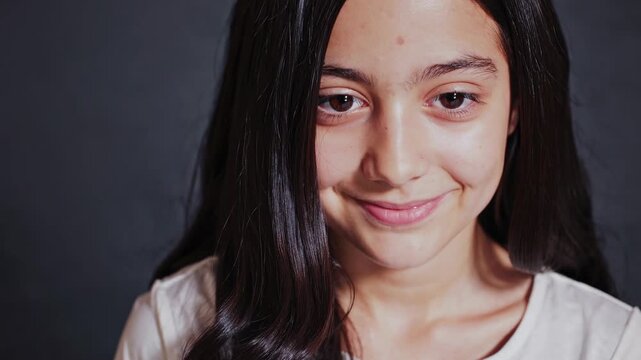 Close-up video shot of a young girl with a neutral expression, capturing her face from a front angle against a dark background, highlighting her features.