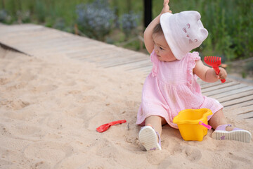 adorable toddler in pink dress plays enthusiastically in a sandy playground with bright yellow bucket and red toy rake under the warm daylight, purple, blue, garden, spring, young, movement, leisure