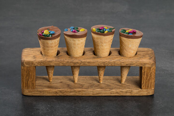 Turkish Waffle dessert. Waffle cones with chocolate nut cream and colored dragees on top. On a wooden stand. Close up. Dark gray background