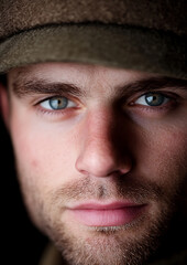 Obraz premium Close-up of a man with striking blue eyes, framed by a cap casting shadows across his face. His gaze is intense, capturing raw emotion, with stubble adding to his rugged demeanor.