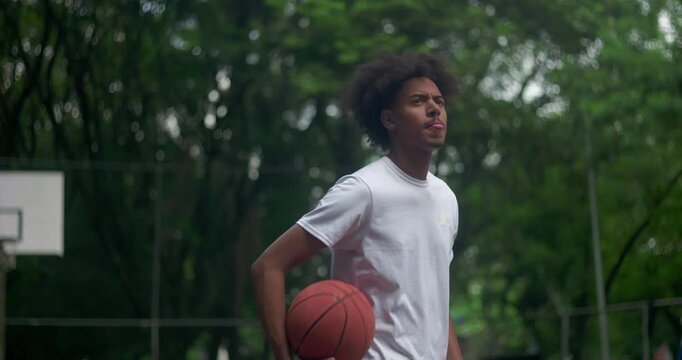 African American man standing on outdoor basketball court holding ball, thoughtful and apprehensive expression, reflecting quietly surrounded by trees