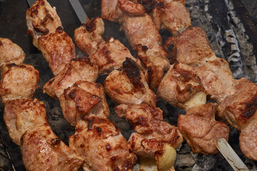 Large pieces of pork being grilled on skewers over hot charcoal. Delicious barbecue with a golden crust cooking outdoors during a picnic or countryside gathering.
