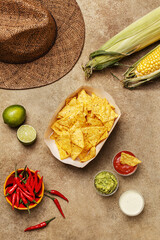 Nachos corn chips with guacamole and salsa dressing, fresh corn cobs, chili peppers, lime and straw hat on beige concrete background. Mexican party snack. Top view, flat lay