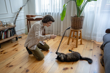 Female pet caretaker playing with cat on floor at home. Woman stimulating hunter instincts with...