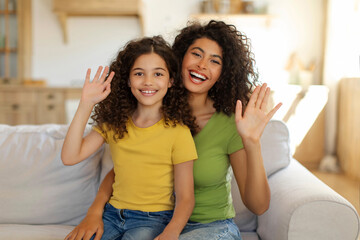 Hello. Happy mom and her preteen daughter waving hands and smiling at camera, sitting on sofa at home. Distance communication concept
