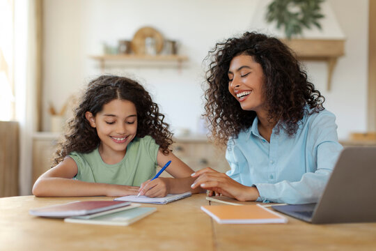Young mother helping daughter with school homework while working on laptop, sitting together at kitchen table. Girl writing in notebook, learn with freelance woman