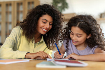 Fototapeta premium Loving mother helping her daughter with school homework, writing in notebook, sitting at their cozy kitchen table at home
