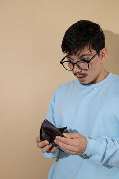 Man with glasses opening his empty wallet with a surprised expression on an isolated background with copy space, concept of lack of money, debt, and personal finance