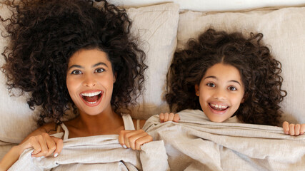Top view of funny daughter lying on bed with young mother and looking at camera, having fun together at lazy weekend at home